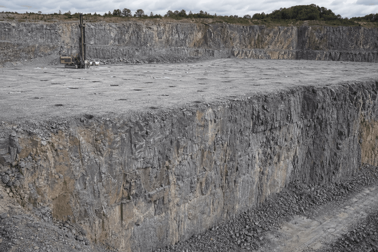 Bancada de cava ou pedreira: malha de furos vista no topo plano da bancada (colares onde a perfuração é feita), com talude vertical ao fundo como frente livre — não furos na face vertical.